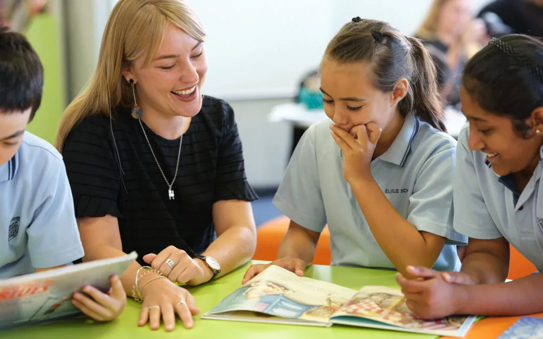 A blonde teacher with a black top laughs while sitting at a table with three primary school students, two girls and one boy, all wearing light blue polo shirts. They are looking at an open book with colorful illustrations. The teacher has a watch and rings on her left hand.