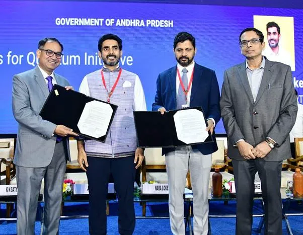 Four men in business attire stand on a stage, two holding signed documents, against a blue backdrop with 'Government of Andhra Pradesh' and 'Nara Lokesh' visible. The two men in the middle, Nara Lokesh and Dr. Pankaj Kumar Dalela, are exchanging the MoU documents.