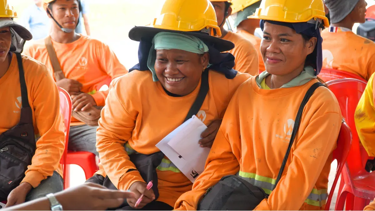 A Lao migrant woman construction worker, Oh, smiles while holding a safety brochure during an onsite training session on labour rights and protection. She is wearing an orange long-sleeved shirt and a yellow hard hat, seated next to another woman, also in a yellow hard hat and orange shirt. Other construction workers are visible in the background.