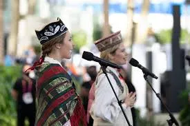 Two women in traditional Latvian folk attire sing into microphones on an outdoor stage. The woman on the left wears a dark cap with white embroidery, a white blouse, and a plaid shawl in red, green, and black. Her hair is in braids with red ribbons. The woman on the right, slightly out of focus, also wears a traditional embroidered top and hat.