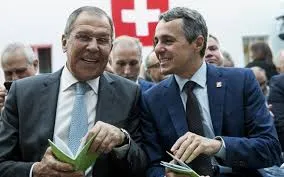 Russian Foreign Minister Sergey Lavrov and Swiss Foreign Minister Ignazio Cassis smiling and holding documents in an indoor setting with a blurred Swiss flag in the background.