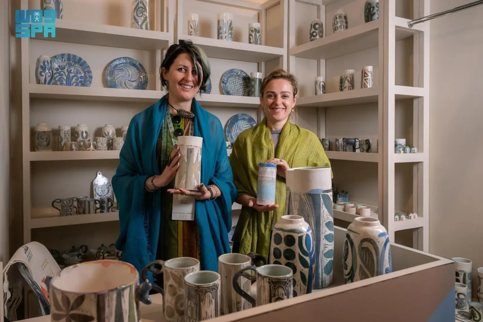 Two Georgian ceramic artists, Anna Gabriadze and Nino Gobaladze, smiling and holding ceramic pieces in their exhibition booth at the Saudi International Handicrafts Week “Banan” in Riyadh, surrounded by their pottery creations.