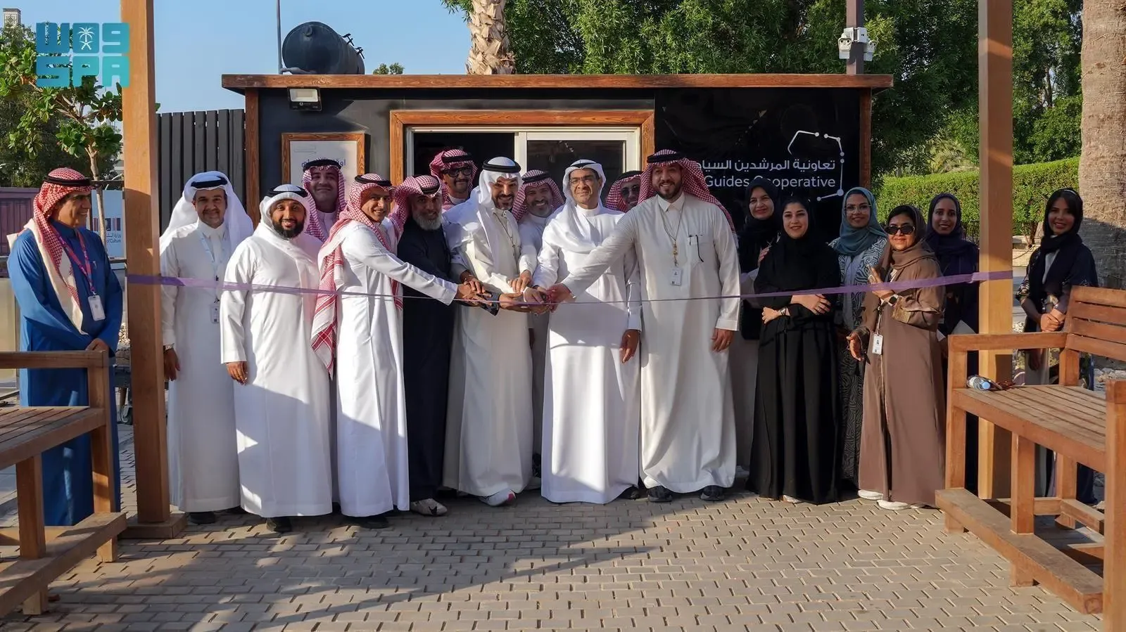A group of men and women in traditional Saudi attire participate in a ribbon-cutting ceremony in front of a modern wooden kiosk with "Guides Cooperative" written on it.