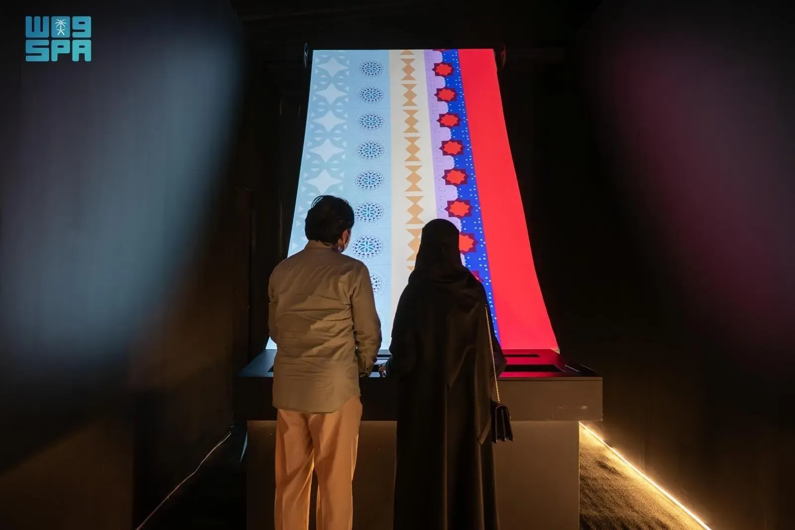Two people, a man and a woman in an abaya, stand with their backs to the viewer, observing a large, illuminated vertical display. The display shows intricate, colorful patterns on both sides, with the left side featuring blue and white designs and the right side showcasing red and blue patterns, suggesting an interactive digital art exhibit.