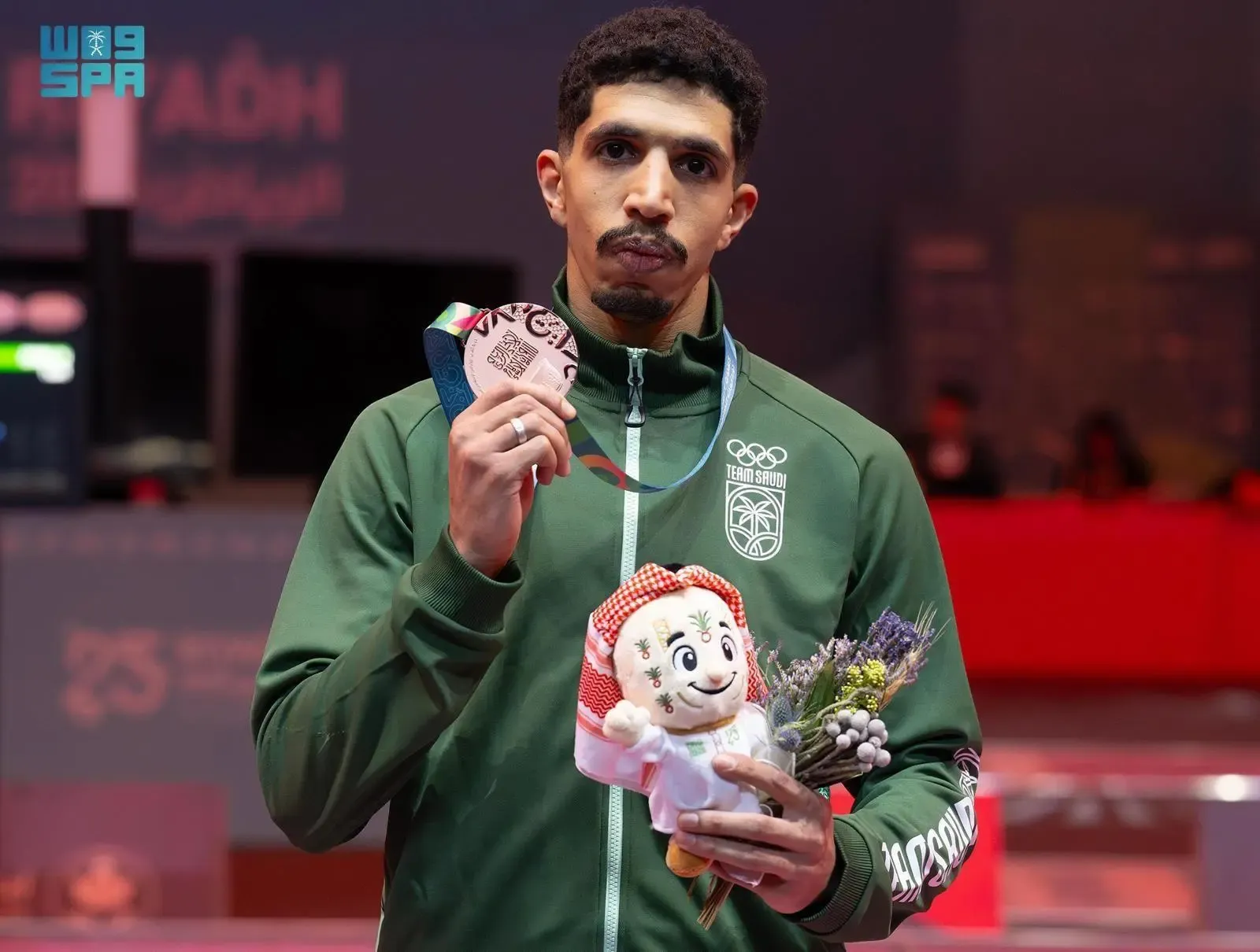 Saudi fencer Khalifa Al-Amiri in a green tracksuit holds a bronze medal and a small plush doll with a bouquet of lavender, looking directly at the camera with a serious expression in a brightly lit indoor arena.
