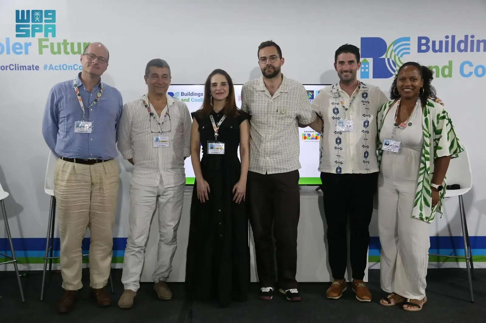 Six individuals, including men and women, standing in a line on a stage in front of a white backdrop with event logos. The participants are diverse in appearance, wearing business casual attire. A screen displaying event branding is visible in the background.