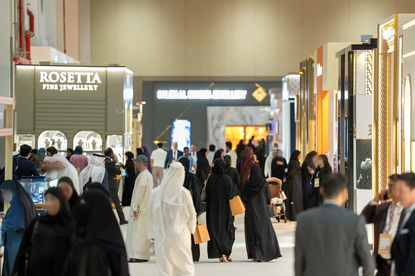 A wide shot of attendees, many in traditional Middle Eastern attire, walking through the Jewellery & Watch Show 2025 exhibition hall with various brand booths like "ROSETTA FINE JEWELLERY" visible.