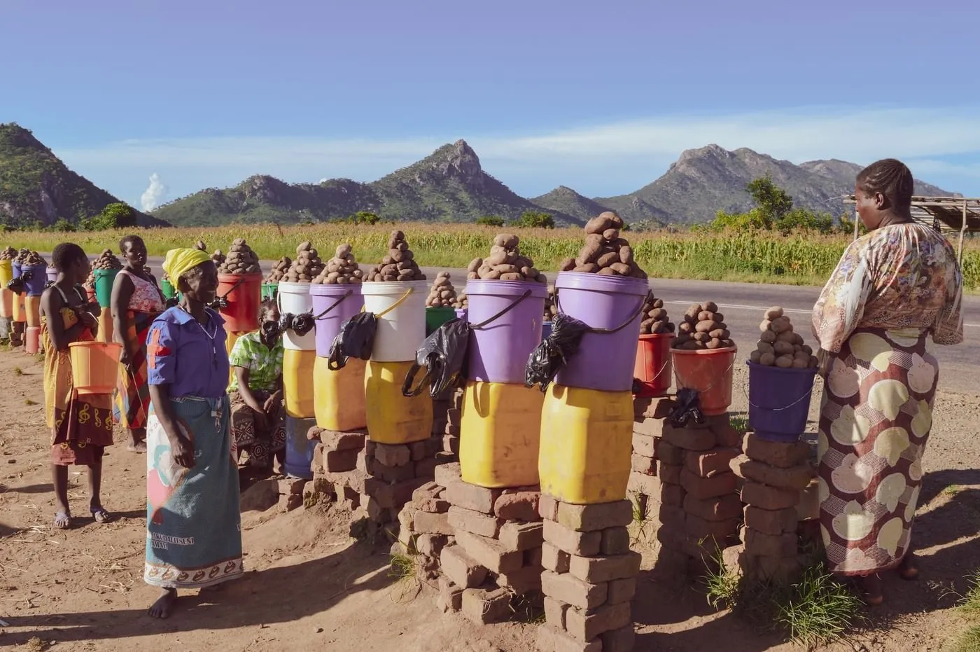 Women stand alongside a rural road in Malawi, selling potatoes from colorful buckets stacked on brick pedestals, with rolling hills and agricultural fields in the background under a clear sky.