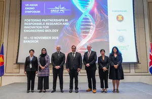 Seven individuals, including men and women, stand formally in a line on a stage in front of a large screen displaying the 'UK-ASEAN ENGBIO SYMPOSIUM' logo, a DNA helix graphic, and text about fostering partnerships in engineering biology research and innovation. The individuals are dressed in business attire, with some wearing suits and others formal dresses. Flags of ASEAN and the UK are visible on either side of the stage.