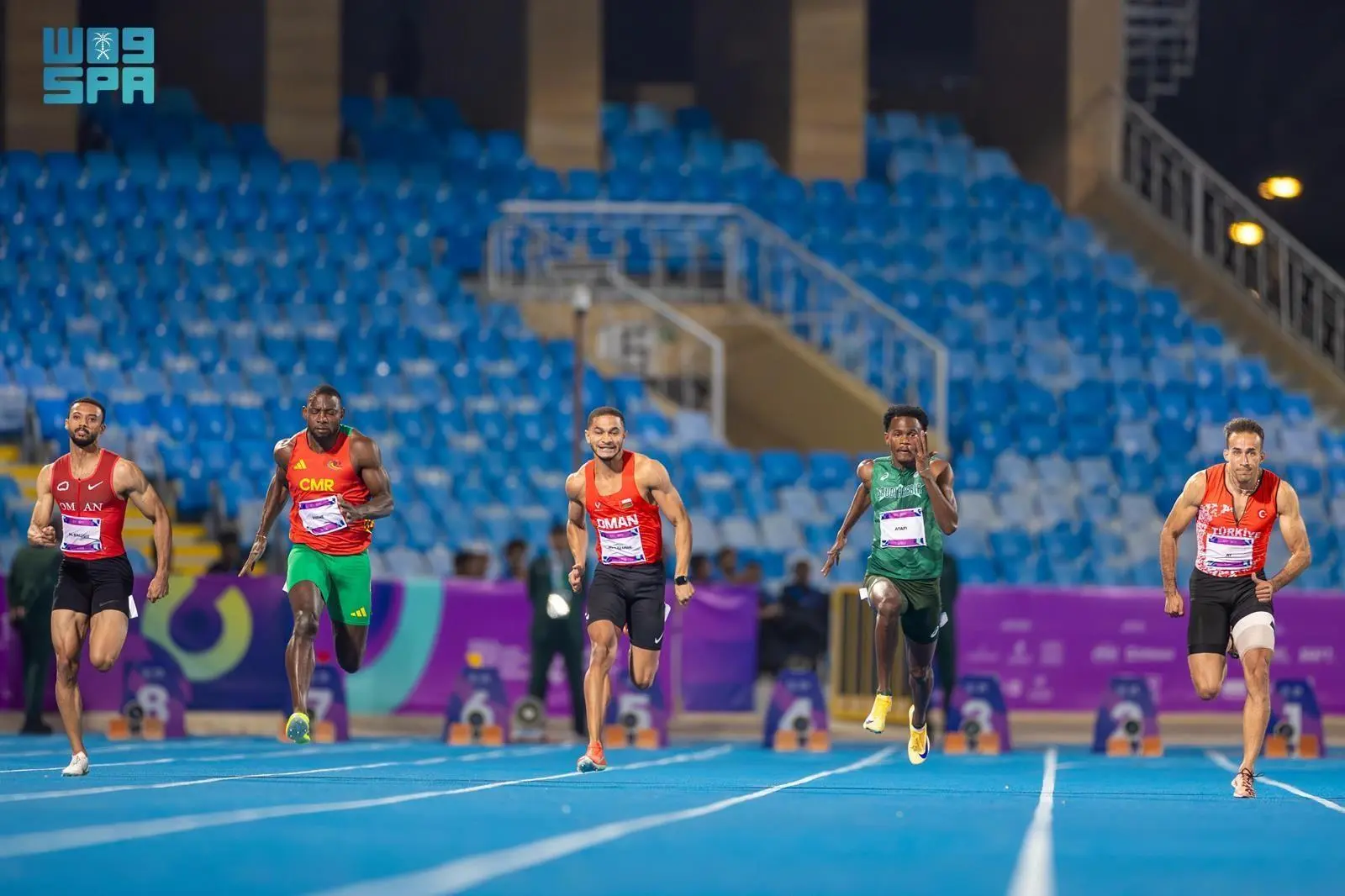 Five male sprinters in various colored athletic wear race on a blue track in a stadium, with empty blue seats in the background during an athletic event.