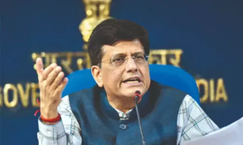 Union Minister Piyush Goyal speaking at a press conference, wearing a blue vest and glasses, with the Government of India emblem in the background.