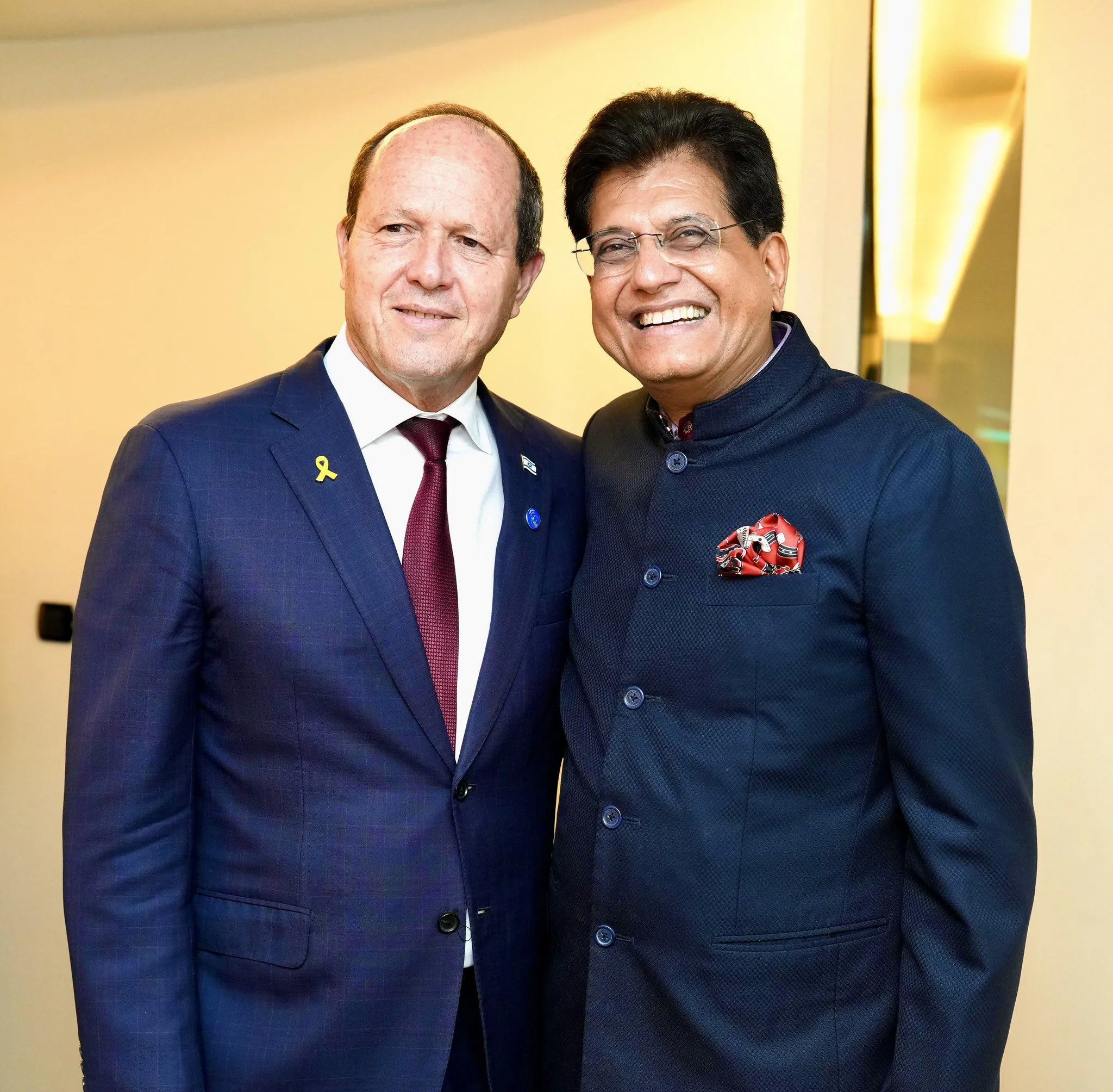Israeli Minister of Economy Nir Barkat and Indian Minister of Commerce Piyush Goyal standing side-by-side; Barkat wears a blue suit with a yellow ribbon pin, while Goyal wears a dark blue Bandhgala and glasses.