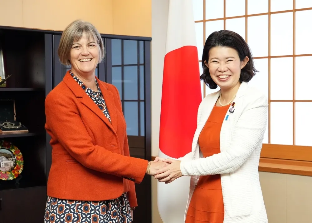 British Ambassador Julia Longbottom (left) shakes hands with Japanese State Minister for Foreign Affairs Dr. Kunimitsu Ayano (right) in front of a Japanese flag during a formal meeting.