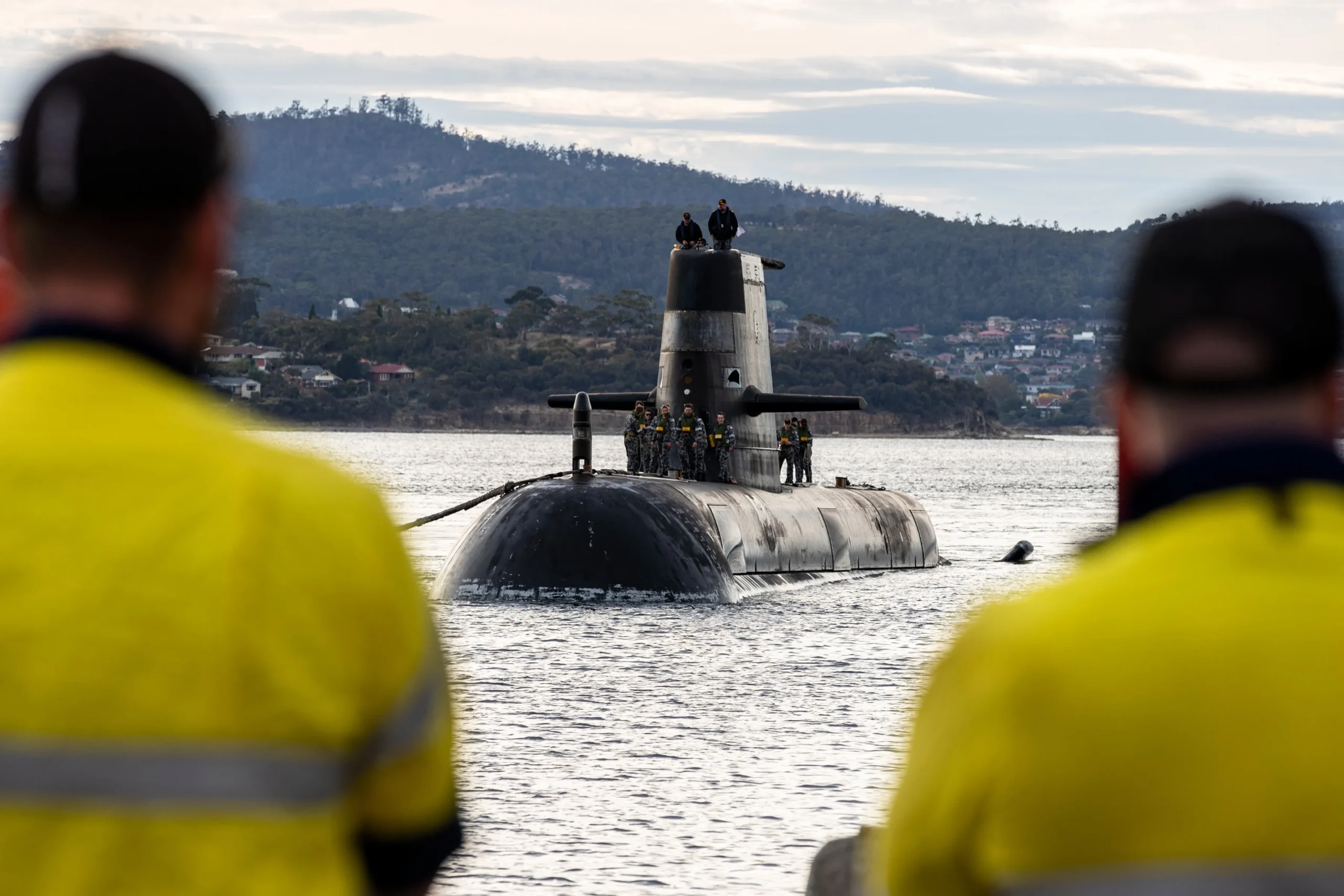 A black Virginia-class submarine floats in the harbor with crew members standing on the conning tower and deck, framed by the blurred shoulders of two workers in yellow high-visibility safety gear in the foreground.