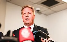 New Zealand government minister Todd McClay speaking into microphones during a press conference, wearing a dark suit and red tie.