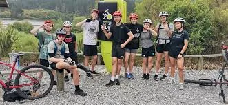 A group of nine young cyclists wearing helmets poses with mountain bikes in front of a black and yellow Great Rides sign on a gravel trail outdoors.