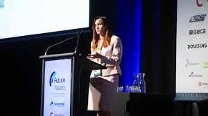 A female speaker with long brown hair, wearing a light pink blazer and dark skirt, gestures while addressing the audience from a podium at the Future Roads 2025 NZ Roading Industry Conference. Sponsor logos including Beca, Leach, and Dowsing are visible on a banner to the right.