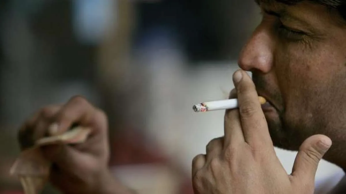 Close-up side profile of a man smoking a cigarette, holding it to his mouth with his right hand against a blurred background.