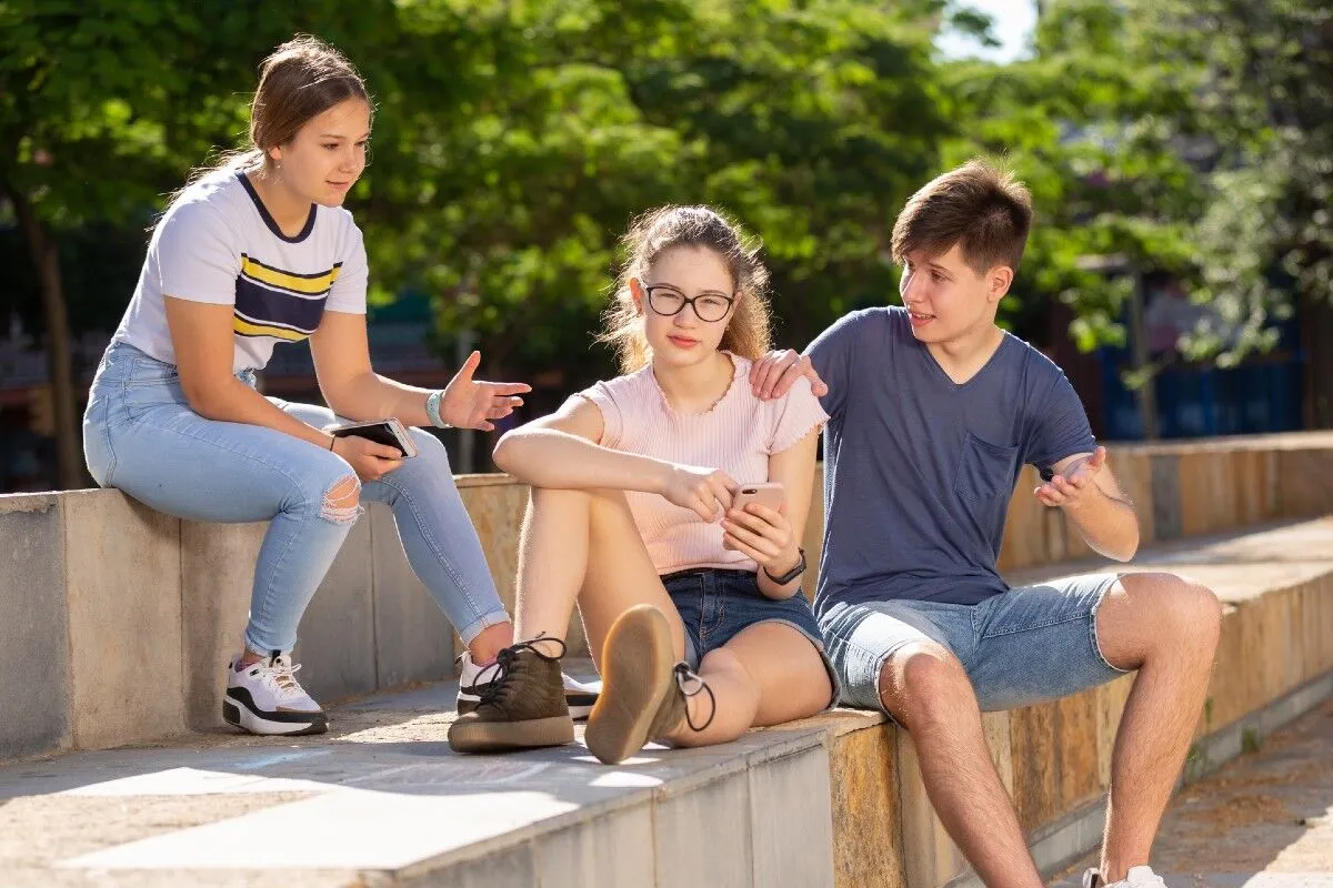 Three teenagers sitting on a concrete ledge outdoors; one girl looks serious and looks away while a boy places a supportive hand on her shoulder and another girl gestures while holding a smartphone.