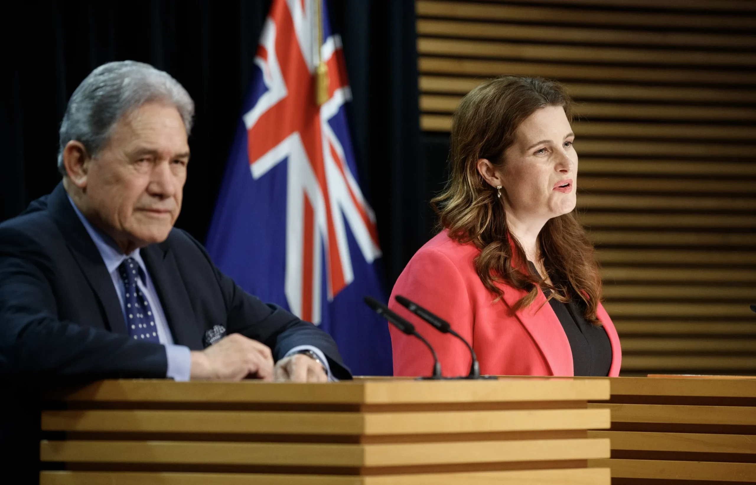 New Zealand Deputy Prime Minister Winston Peters and Finance Minister Nicola Willis standing at podiums during a press conference regarding the Interislander ferry replacement project, with the New Zealand flag in the background.