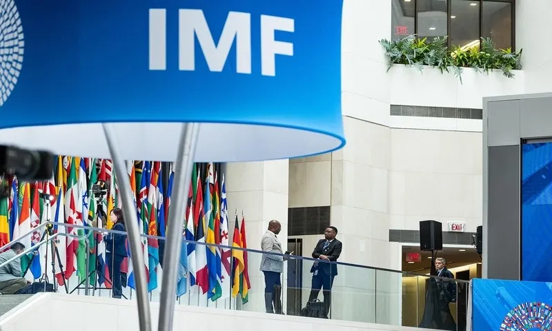 A large blue IMF logo sign hangs in a modern atrium in Washington DC, with international flags and officials conversing on a balcony in the background.