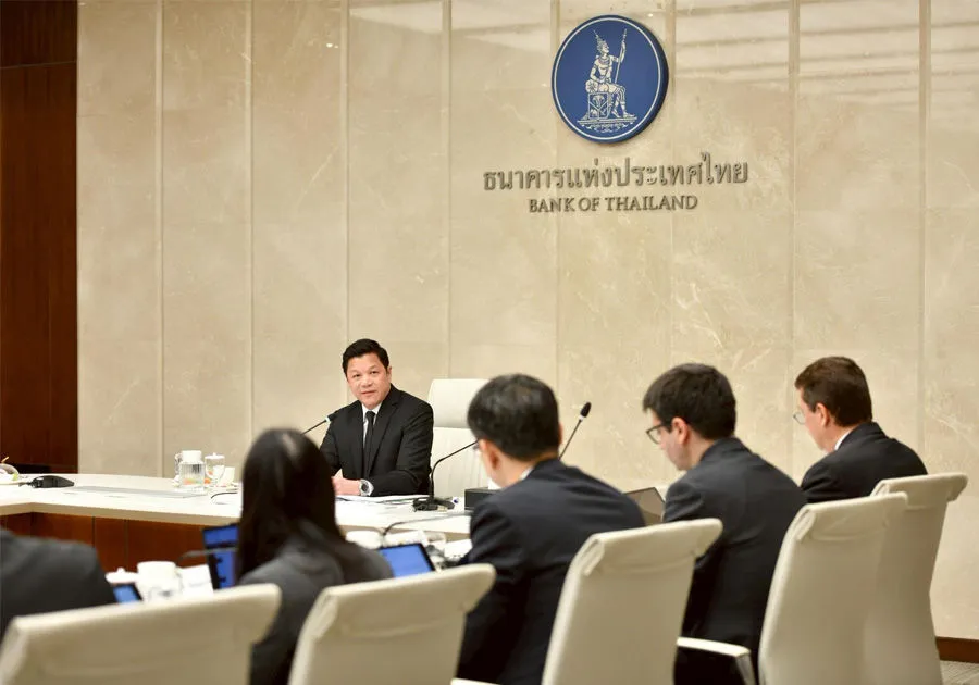 Bank of Thailand Governor Sethaput Suthiwartnarueput speaking at the head of a conference table during a meeting, with the Bank of Thailand logo and Thai script visible on the beige marble wall behind him.