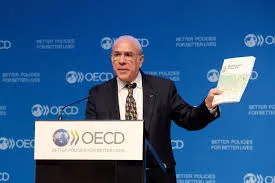 Former OECD Secretary-General Angel Gurría standing at a podium holding a white report against a blue background with OECD logos.