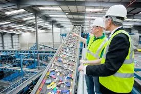 Two men wearing hard hats and high-visibility safety vests stand on an elevated walkway, inspecting a conveyor belt transporting mixed plastic waste at an industrial recycling facility.