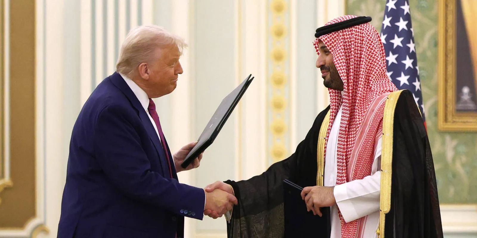 Former US President Donald Trump shakes hands with Saudi Crown Prince Mohammed bin Salman while holding document folders during a bilateral meeting, flanked by US and Saudi flags.