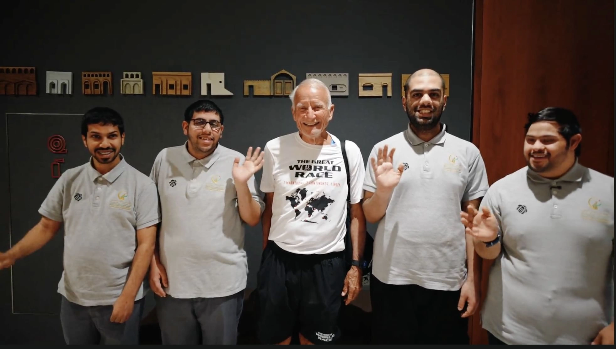 A smiling senior runner wearing a 'The Great World Race' t-shirt stands center, flanked by four young men from the Zayed Authority for People of Determination wearing matching grey polo shirts, waving at the camera against a dark wall featuring architectural models in Abu Dhabi.