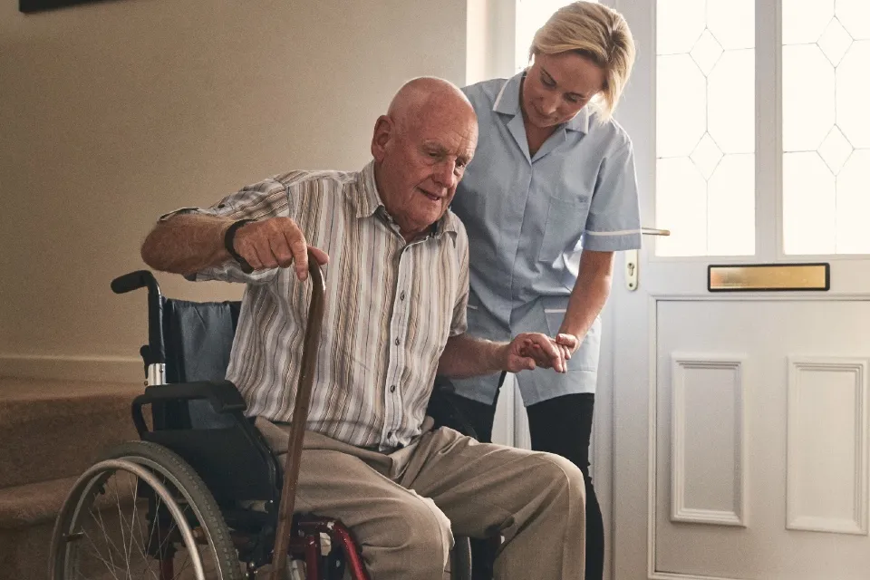 A uniformed female caregiver gently holds the hand of an elderly man sitting in a wheelchair and holding a wooden walking cane near a doorway.