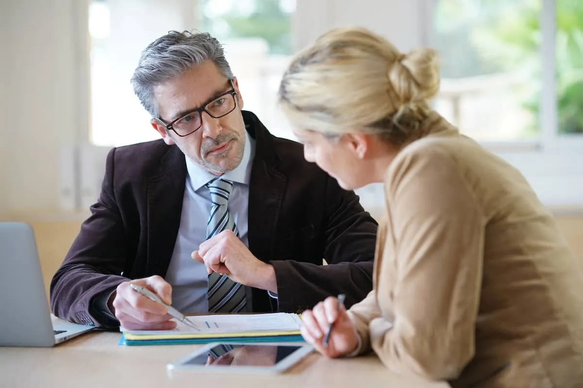 A male legal professional in a suit and glasses reviews a document with a female client at a desk, representing housing loss prevention advice.