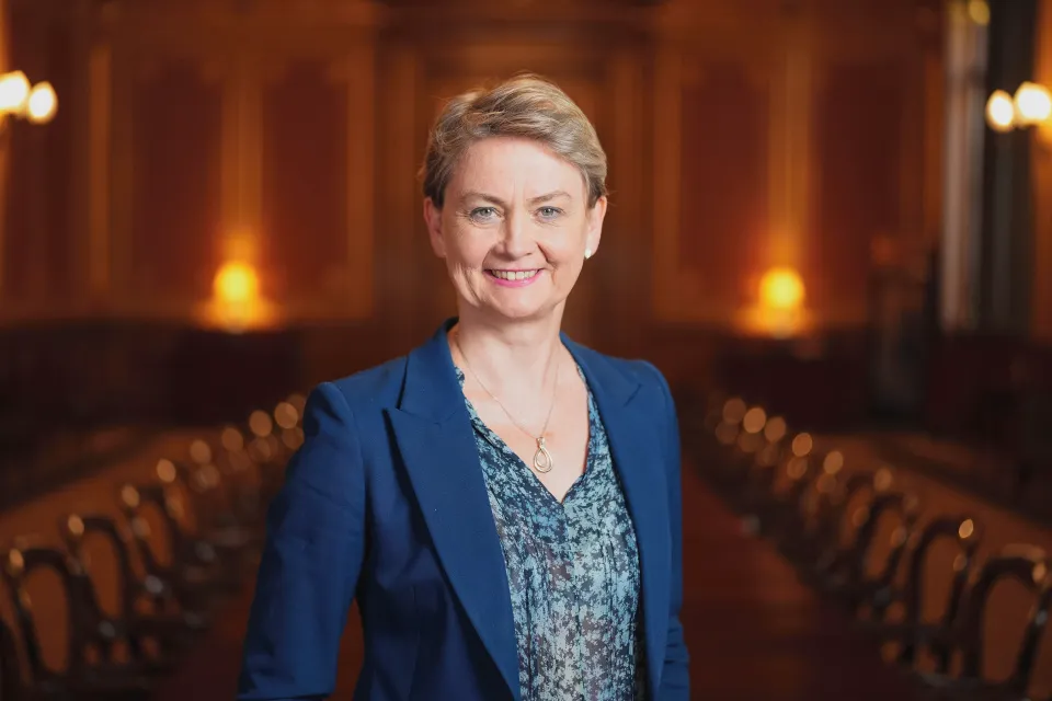 A portrait of UK Foreign Secretary Yvette Cooper smiling, wearing a blue blazer and patterned top, standing in a formal wood-paneled room with rows of wooden chairs visible in the blurred background.