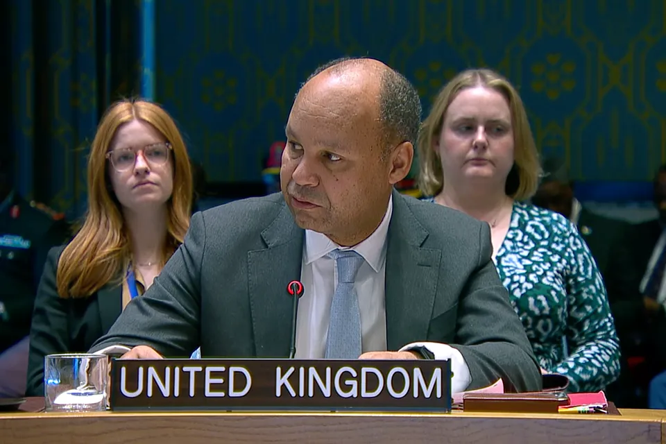 UK Chargé d’Affaires Ambassador James Kariuki speaks into a microphone behind a United Kingdom nameplate at a UN Security Council meeting, with two colleagues seated behind him against a blue patterned wall.