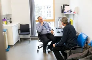 A male doctor sits at a desk in a clinical consultation room, listening intently to a male patient seated opposite him during a medical appointment.