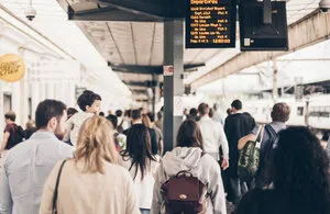 Passengers walking along a busy train station platform with a digital departure board hanging overhead and an Upper Crust sign visible on the left.