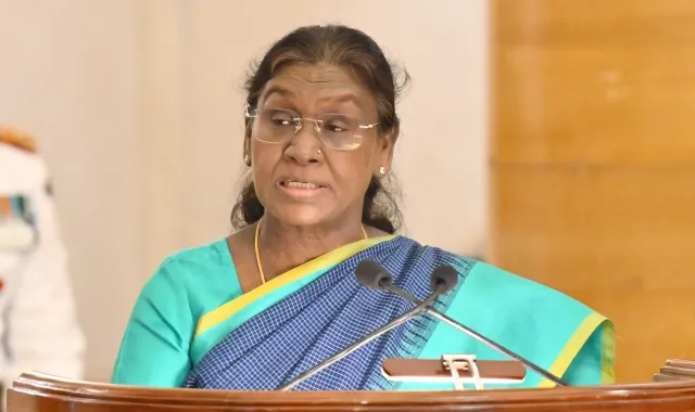 President of India Droupadi Murmu delivering a speech at a podium, wearing a teal saree with a checkered pattern and glasses, with microphones visible in the foreground.