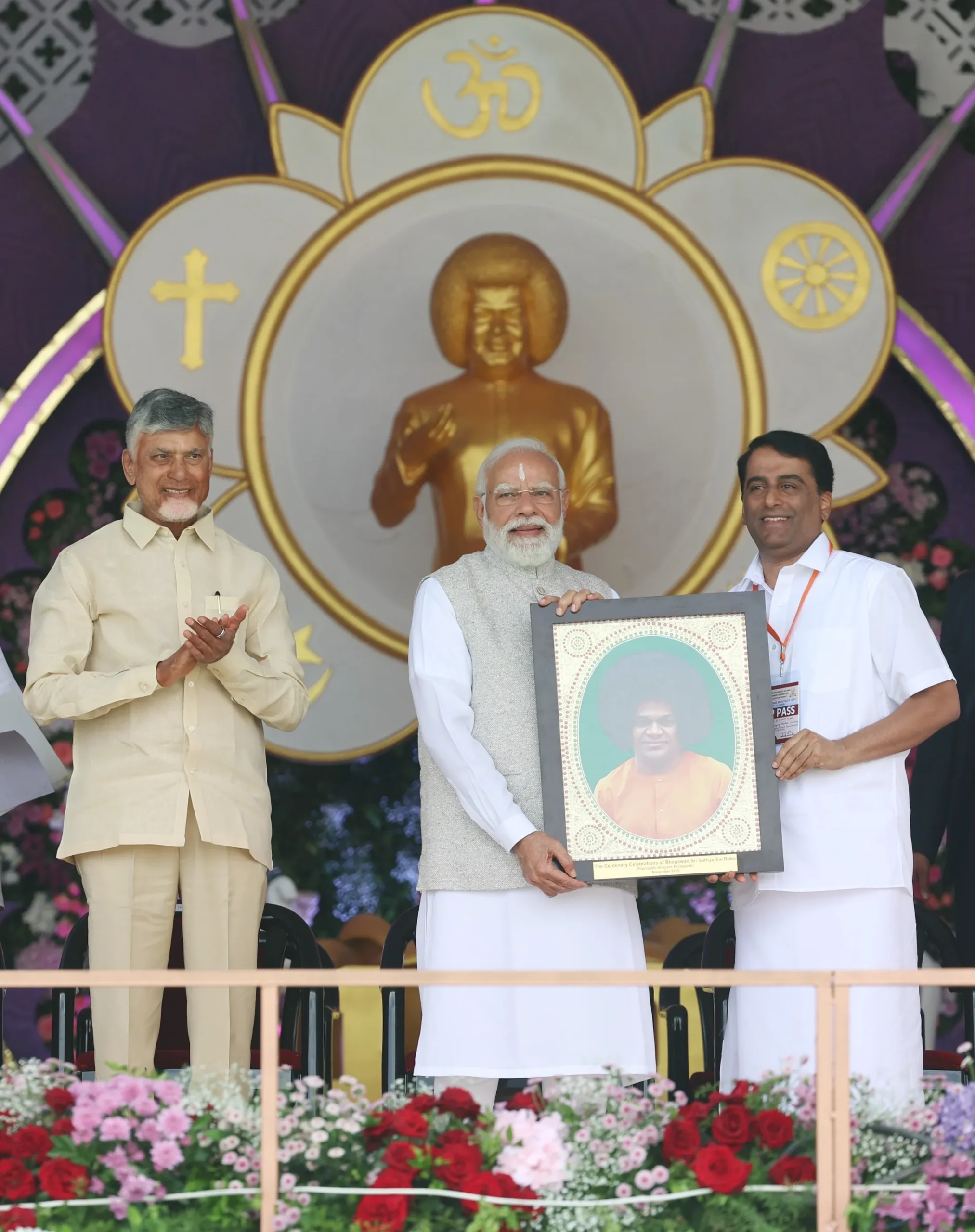 Prime Minister Narendra Modi holds a framed portrait of Sri Sathya Sai Baba on stage in Puttaparthi, flanked by Andhra Pradesh CM N. Chandrababu Naidu applauding on the left and trustee RJ Rathnakar on the right, with a golden statue in the background.