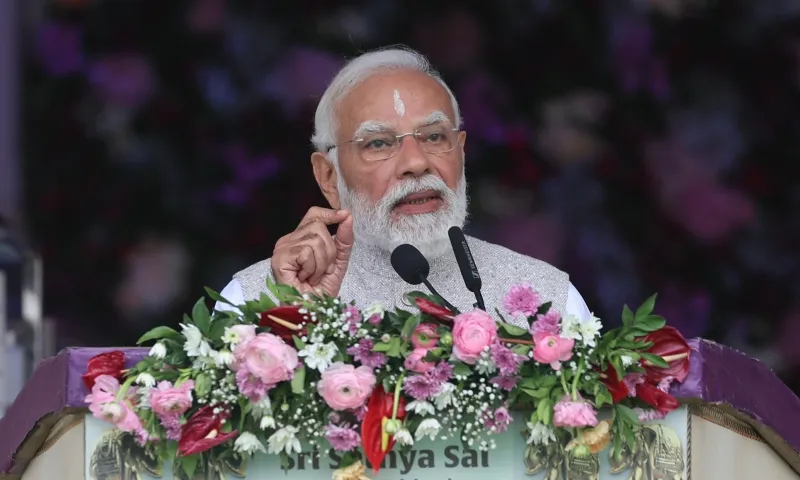 Prime Minister Narendra Modi speaking at a podium adorned with pink and white flowers during the Sri Sathya Sai Baba birth centenary celebrations in Puttaparthi, Andhra Pradesh. He is wearing a light grey vest over a white kurta and has a tilak on his forehead