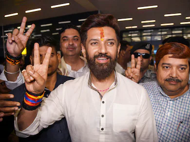 Chirag Paswan smiling and flashing a victory sign with a tilak on his forehead while surrounded by supporters and security personnel.