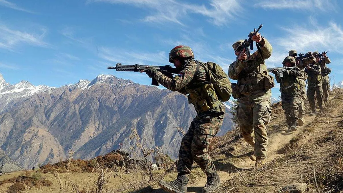 Indian Army soldiers equipped with rifles and wearing the new digitally printed combat coats patrolling in rugged mountainous terrain with snow-capped peaks in the background.