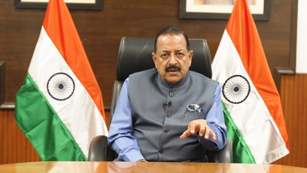 Union Minister Dr. Jitendra Singh seated at a wooden desk, wearing a grey Nehru jacket over a blue shirt, gesturing with his left hand while speaking, flanked by two Indian national flags in an office setting.