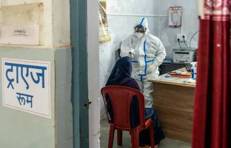 A healthcare worker wearing a full white PPE suit and face mask measures the temperature of a seated woman in a blue headscarf using a thermal scanner inside a clinic room labeled "Triage Room" in Hindi.