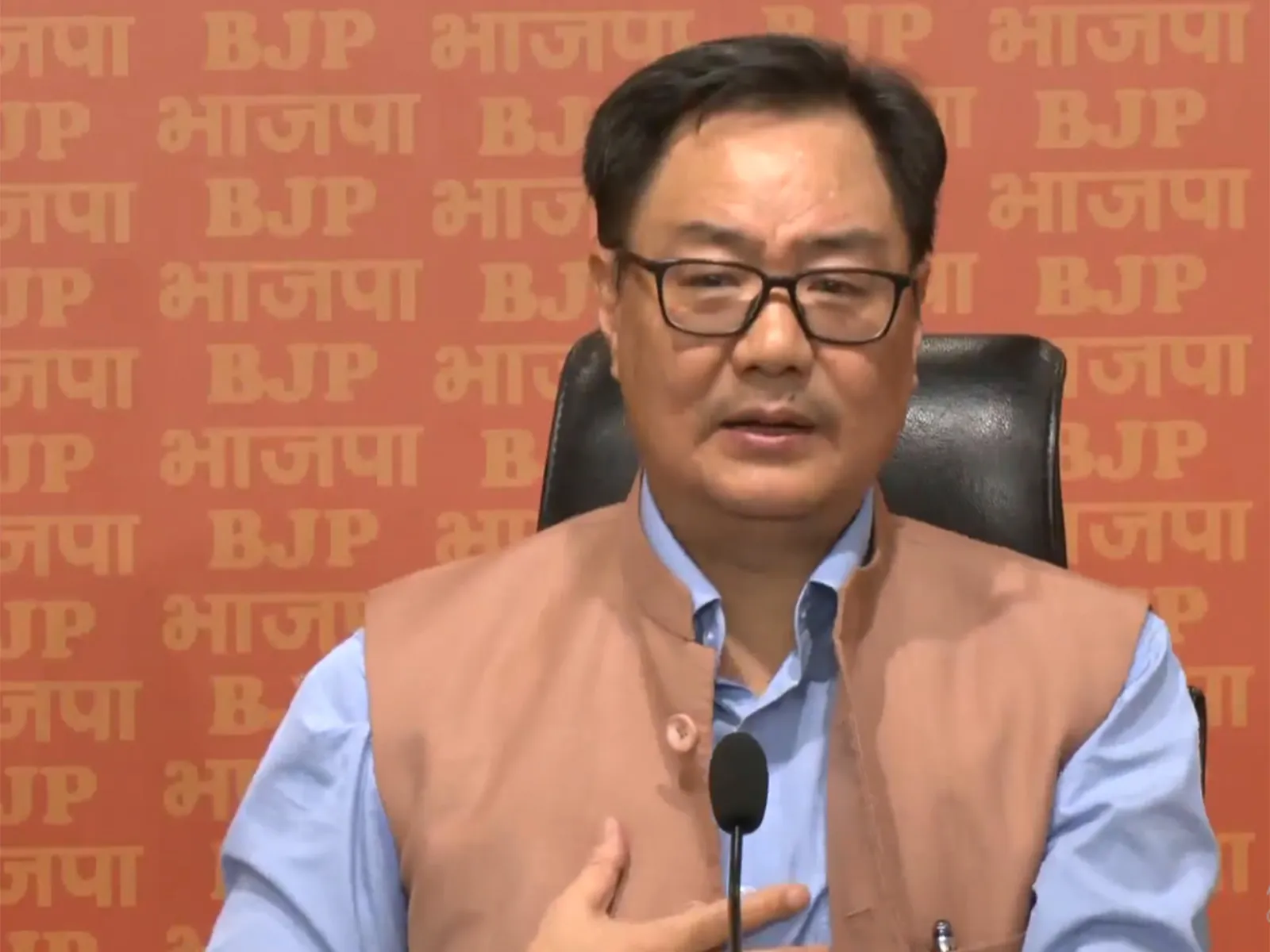 Union Minister Kiren Rijiju speaking at a press conference, wearing a beige Nehru jacket and glasses, against an orange background featuring the BJP logo.