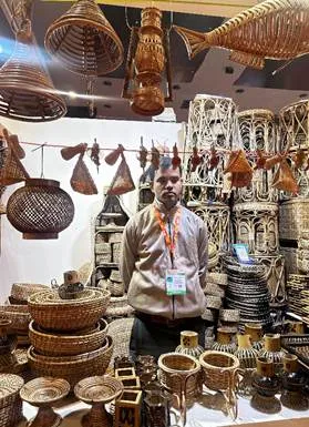 A male artisan wearing an ID lanyard stands inside a stall filled with woven cane and bamboo products, including hanging fish-shaped lamps, round lanterns, and stacked baskets at the India International Trade Fair. 