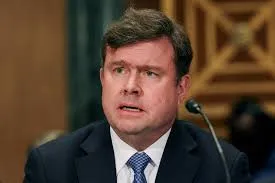 U.S. Senator Todd Young wearing a dark suit and blue tie speaking into a microphone during a committee hearing with a blurred background.