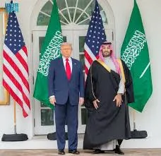 Donald Trump and Saudi Crown Prince Mohammed bin Salman stand side-by-side in a formal diplomatic setting, flanked by the national flags of the United States and Saudi Arabia.
