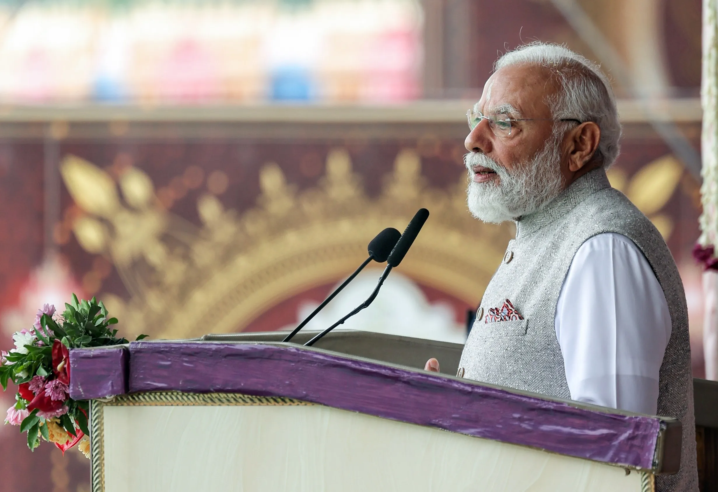 Indian Prime Minister Narendra Modi speaking at a podium wearing a grey vest and glasses during a formal event.