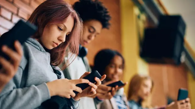 Group of diverse teenagers leaning against a brick wall at school, looking down at smartphones in their hands.