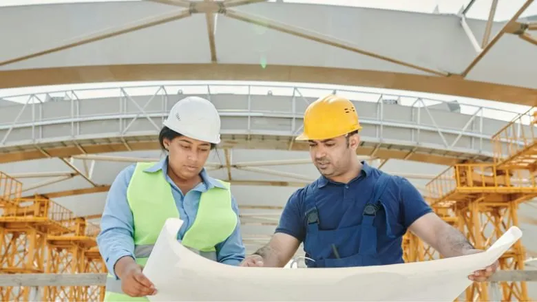 A female construction worker wearing a white hard hat and green safety vest reviews a large blueprint with a male colleague in a yellow hard hat and blue overalls at an industrial construction site.